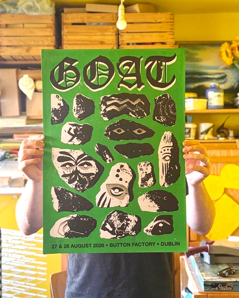 A person holds up a green concert poster for the band Goat, featuring black and beige abstract rock art and text reading 'GOAT 27 & 28 AUGUST 2026 • BUTTON FACTORY • DUBLIN'. The background shows a workshop or studio with wooden crates and shelves.