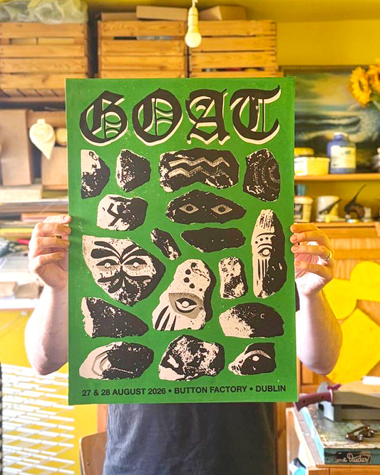 A person holds up a green concert poster for the band Goat, featuring black and beige abstract rock art and text reading 'GOAT 27 & 28 AUGUST 2026 • BUTTON FACTORY • DUBLIN'. The background shows a workshop or studio with wooden crates and shelves.